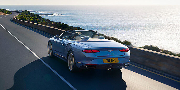 Bentley Kuwait New Bentley Continental GTC Azure convertible rear view in Light Windsor blue paint with 22 inch ten spoke wheels driving dynamically on an ocean road in the sunshine