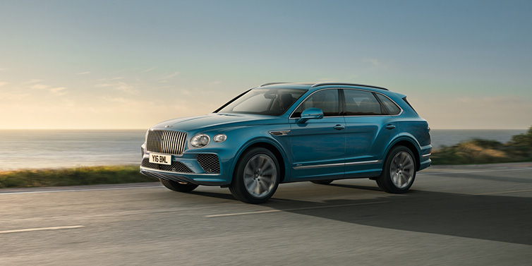 Bentley Kuwait Bentley Bentayga Azure SUV in Topaz blue paint driving dynamically by the ocean with 22 inch 10 spoke directional wheels