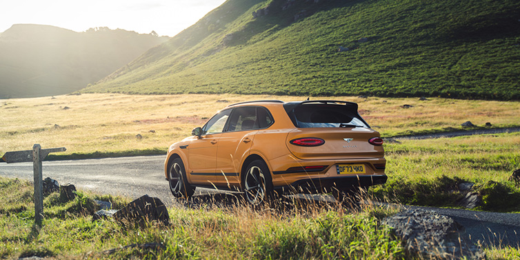 Bentley Kuwait Bentley Bentayga S SUV rear three quarter in Sunburst Gold paint on a road surrounded by green hills