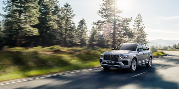 Bentley Kuwait Bentley Bentayga SUV front three quarter in Moonbeam silver paint driving dynamically along a tree-lined road