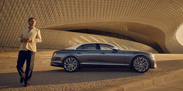 Bentley Kuwait Bentley Flying Spur Mulliner sedan in Rose Gold over Magnetic duotone paint parked by a curved wall with person in suit jacket standing on the left